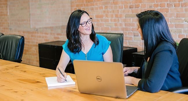 Zwei Frauen arbeiten gemeinsam an einem Tisch vor Ihnen liegt ein Laptop.