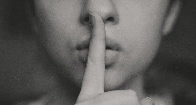 Eine Frau mit der ausgestreckten Zeigefinger mit der Fingerspitze zur Nase zeigend senkrecht auf den eigenen Lippen. Bitte um Ruhe oder Verschwiegenheit.