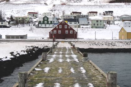 Photo by Luciano Braga Ein Steg mit rotem Haus vor einer verschneiten Landschaft an einem See in Island.